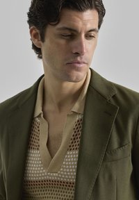 Blazer verde oliva sobre un suéter de punto beige con escote en V y patrones geométricos en rojo, blanco y verde. Textura suave, corte ajustado.