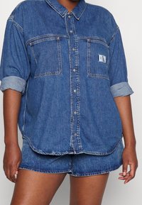 Denimskjorta, ljusblå, med knäppning fram, två bröstfickor, uppvikta ärmar och CK-logotyp. Matchas med passande denimshorts.