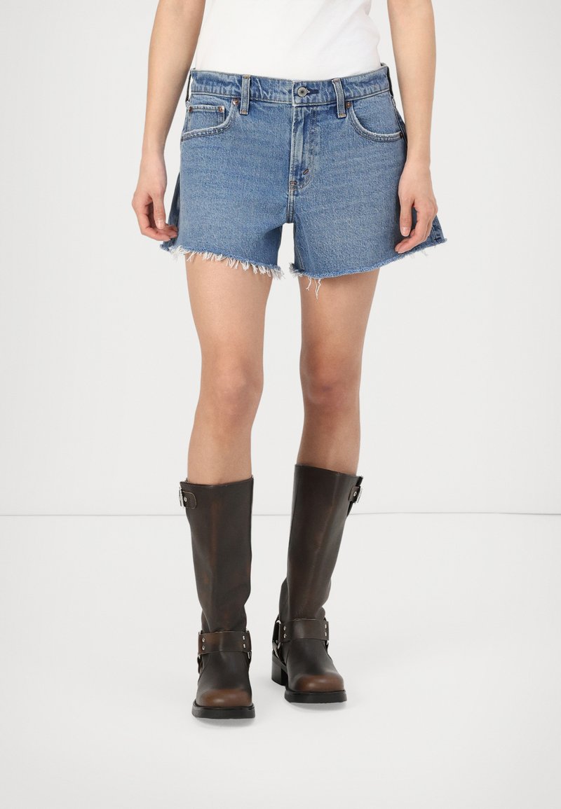 Lichtblauwe denimshorts met franjes aan de hem, gecombineerd met hoge, donkerbruine leren laarzen met gespen en textuurdetails.
