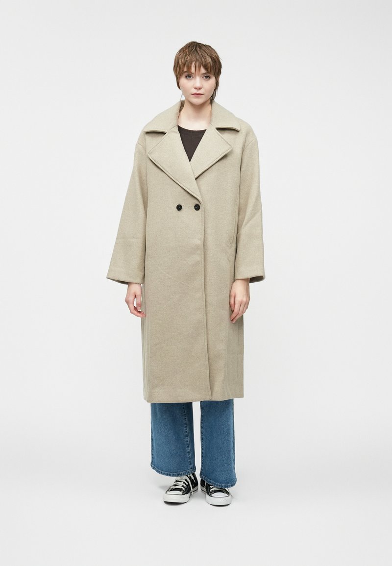 Μπεζ μάλλινο παλτό με φαρδύ γιακά, διπλό κούμπωμα και χαλαρή εφαρμογή. Συνδυασμένο με φλαρντ τζιν παντελόνι και μαύρα αθλητικά παπούτσια.