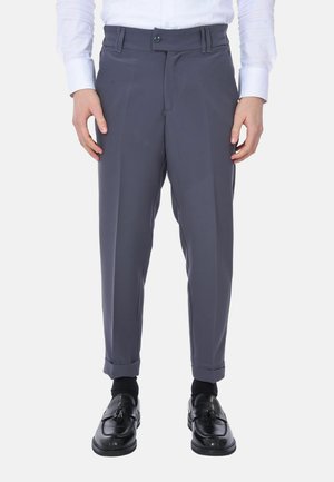 Uomo con pantaloni formali grigi, calzini neri e mocassini neri con nappine, con camicia bianca infilata, in piedi davanti a uno sfondo semplice.