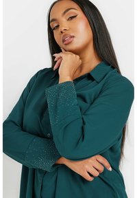 Camicia color verde acqua con colletto a punta e chiusura a bottoni, caratterizzata da una texture liscia e decorazioni brillanti vicino ai polsini.