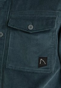 Dark teal corduroy shirt featuring a buttoned chest pocket and a black label with a white graphic. Textured surface with vertical ridges.
