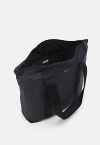 Mustasta kestävästä kankaasta valmistettu kangaskassi, jossa on vetoketjullinen yläosa, säädettävät hihnat ja pieni sisätasku. Yksinkertainen muotoilu logolla.