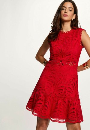 Donna che indossa un vestito rosso senza maniche in pizzo con motivi floreali e bordi smerlati, accessoriata con braccialetti d'oro e orecchini a cerchio.