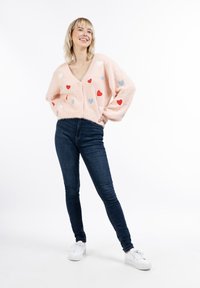 Rózsaszín, bolyhos kardigán, sokszínű szívecske mintákkal, mély V-kivágással és gombos záródással, sötét, skinny farmernadrággal és fehér sportcipővel párosítva.