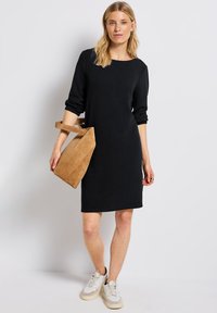 Robe noire en tricot avec des manches trois-quarts, un col rond et une coupe droite. Associée à un sac à main en daim beige et des baskets blanches.