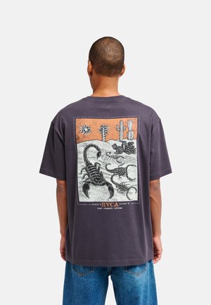 Persoon met kort haar die een donker T-shirt draagt met een woestijnscène met schorpioenen, cactussen en een zon op de rug.