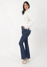 Pull-over blanc à côtes avec col zippé, associé à un jean flare bleu foncé et des sandales à talons aiguilles blanches.