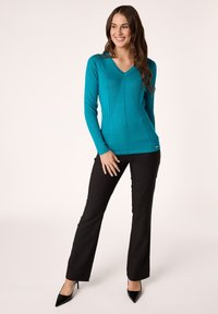 Maglia lunga teal con scollo a V e design a coste testurizzate, abbinata a pantaloni neri sartoriali e décolleté nere a punta.