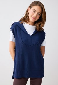 Jeune femme aux cheveux bruns mi-longs, portant un pull sans manches en maille bleu marine par-dessus une chemise à manches courtes blanche.