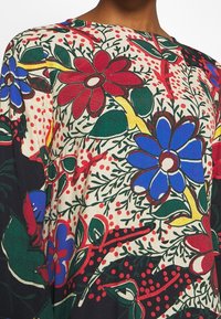 Veelgekleurde blouse met een bloemenpatroon van rode, blauwe en groene bloemen op een beige achtergrond, gemaakt van textuurstof.