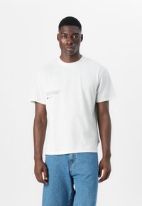 Weißes Baumwoll-T-Shirt mit Rundhalsausschnitt, kurzen Ärmeln und einem kleinen Druckdetail auf der linken Seite. Kombiniert mit blauen Jeans.
