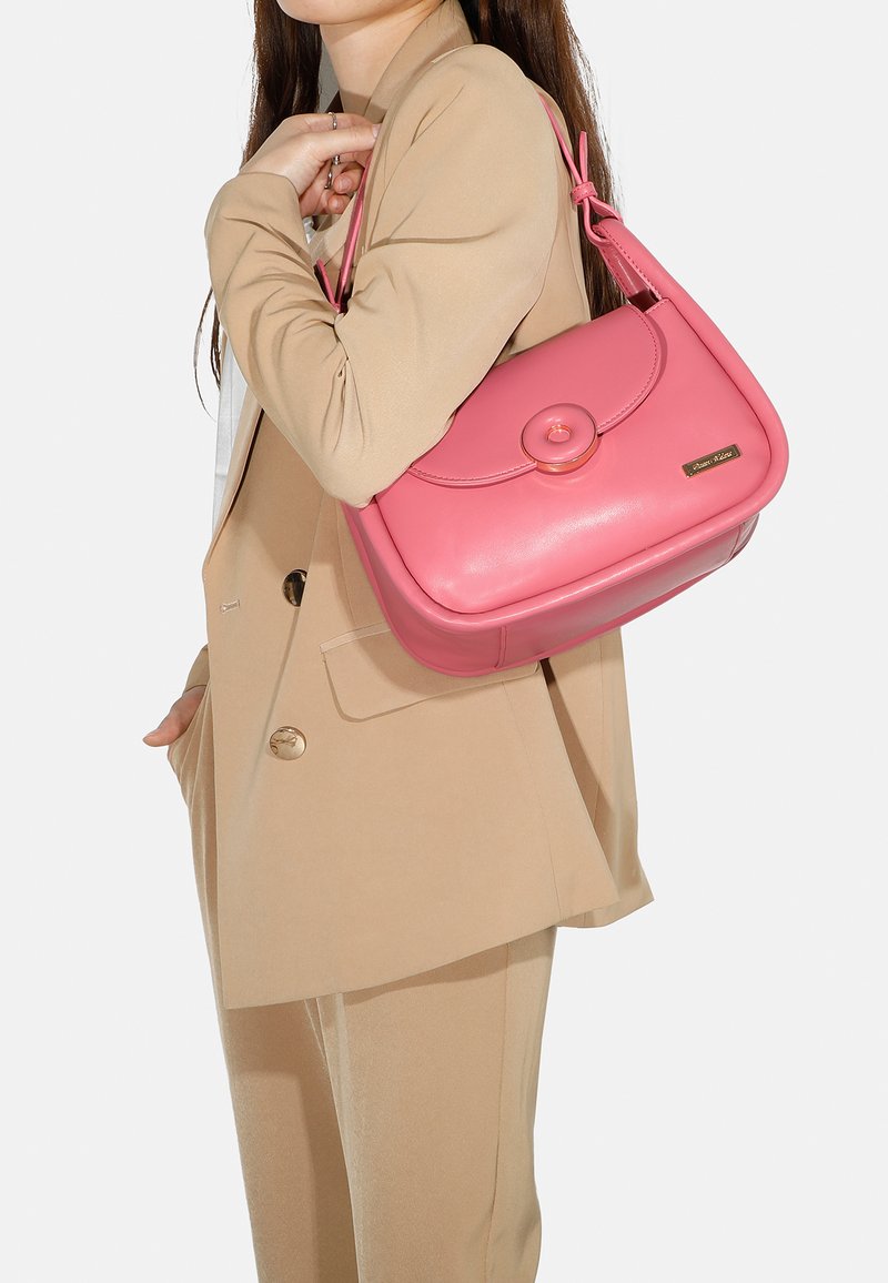 Borsa a tracolla in pelle rosa con una pattina arrotondata, un grande bottone decorativo e una tracolla, mostrata su un modello che indossa un completo beige.