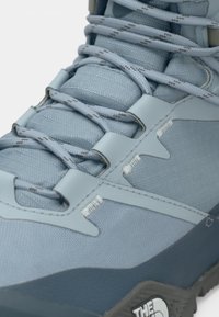 Svijetloplata planinarska cipela s teksturiranim gornjim dijelom od sintetičkog materijala, ojačanim otvorima za pertle, kontrastnim tamnoplavim potplatom i brendiranim detaljem sa strane.