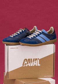 Paire de baskets en daim bleu avec des bandes bleu clair et des semelles beiges sur des boîtes à chaussures beiges et blanches empilées sur un fond rouge.