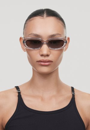 Sluneční brýle s průhledným rámem, tmavými skly a úzkým obdélníkovým tvarem. Nosí je osoba v černém tílku s minimálními detaily.