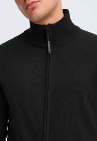 Svart zip-jakke med høy krage, ribbet tekstur og glidelås med logo-kort. Enkelt design uten tilleggsmønstre eller -detaljer.