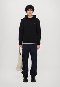Black hooded sweatshirt, navy trousers, black shoes, holding a beige tote bag. Simple design with a small logo on the chest.
