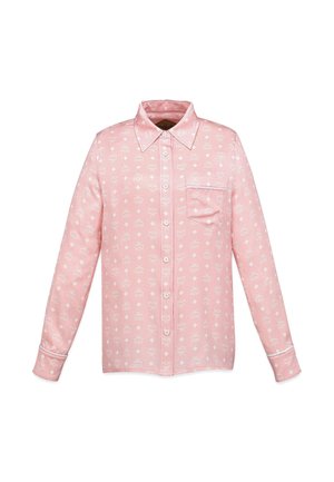 Kvinde med langt hår iført matchende lyserødt mønstret pyjamasæt med skjorte og bukser samt hvide sneakers mod en lys grå baggrund.