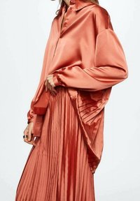 Satin langærmet skjorte i koralfarve med knaplukning og en afslappet pasform, parret med en plisseret koralskørter, begge med glatte teksturer.