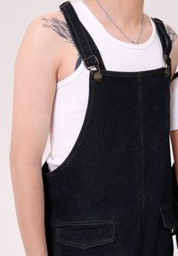 Denim kombinezoni temno modre barve z nastavljivimi naramnicami, kovinsko opremo in dvema sprednjima žepoma. Obuti preko bele majice brez rokavov.