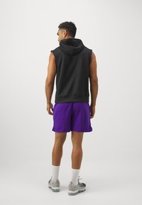 Cappuccio senza maniche nero, pantaloni corti viola, design atletico. Scarpe grigie con dettagli gialli, calze bianche. Vista posteriore su uno sfondo semplice.