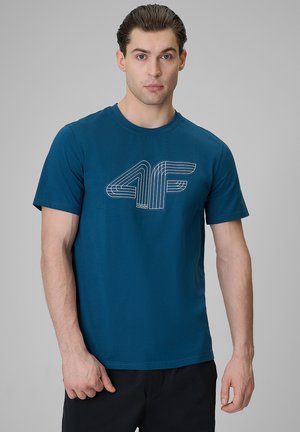 Mężczyzna o krótkich brązowych włosach, ubrany w morski t-shirt z krótkim rękawem z białym obrysowanym logo "4F" na piersi, stoi na szarym tle.