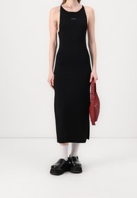 Robe maxi noire ajustée avec un décolleté rond et une fente latérale, associée à un sac à main rouge et des chaussures plateformes noires épaisses. Chaussettes blanches visibles.