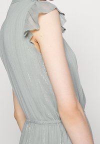 Närbild av en person som bär en ljusgrå ärmlös klänning med subtila vertikala ränder och volangdetaljer på axlarna.