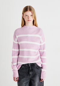 Rosa og hvitstripet strikkegenser med høy hals, ribbestruktur og lange, litt oversized ermer, sammen med grå jeans.