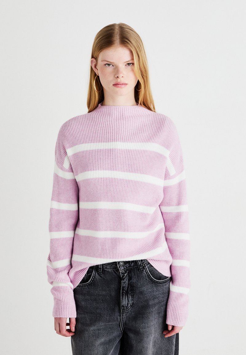 Rosa og hvitstripet strikkegenser med høy hals, ribbestruktur og lange, litt oversized ermer, sammen med grå jeans.