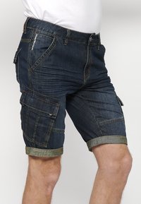 Homme portant un short cargo en denim bleu foncé avec des revers roulés et plusieurs poches, associé à une chemise blanche.