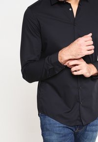 Black long-sleeve shirt with a button-down collar and smooth texture. Features a fitted design and subtle front buttons. Worn with denim.