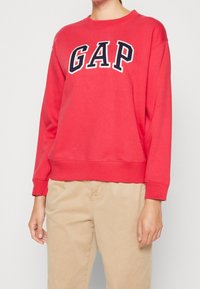 Czerwona bluza z kapturem z logo "GAP" w białym i granatowym. Okrągły dekolt, długie rękawy, ściągacze na rękawach i na dole. W zestawieniu z beżowymi spodniami.