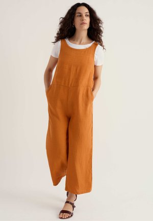 Mujer con cabello rizado caminando hacia adelante, vistiendo un mono sin mangas naranja sobre una camiseta blanca de manga corta, con las manos en los bolsillos y sandalias.