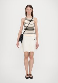 Striped tank top in beige and black, paired with a white pleated skirt. Model carries a black fringe crossbody bag. Flat black shoes.