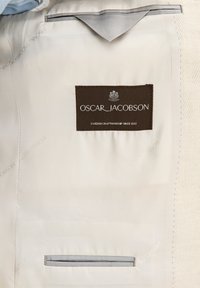 Beige tyg med en slät textur, med en mörkbrun etikett som läser "OSCAR_JACOBSON" och en horisontell ficka med grå kant.