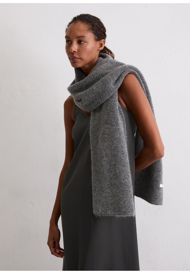 Écharpe en laine grise enroulée autour du cou, présentant une texture douce et un bord effiloché. Le modèle porte une robe sombre sans manches.