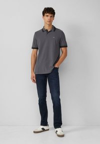 Grijze polo met zwarte accenten, gecombineerd met donkerblauwe jeans. Model draagt witte sneakers met donkere strepen. Eenvoudig, sportief ontwerp.