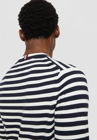 Sweater riscado em azul-marinho e branco com um pescoço redondo canelado. Acento vermelho notável no pescoço, textura de malha suave e design ajustado.