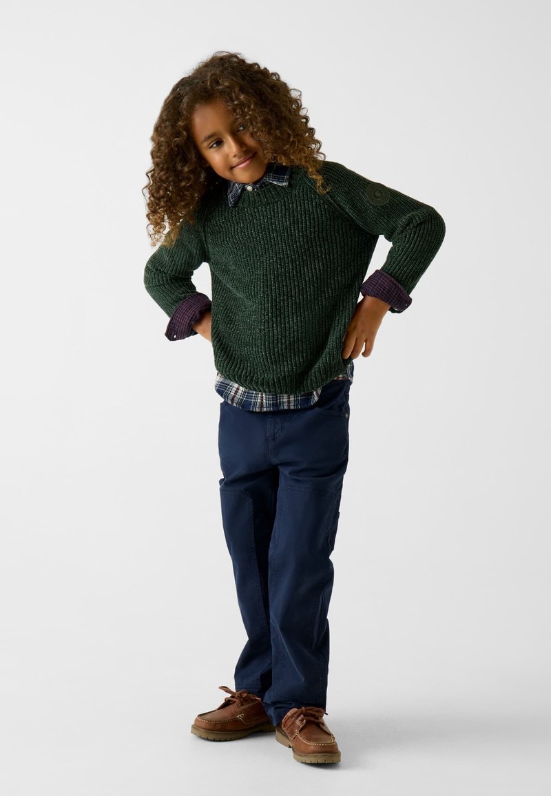 Maglione verde a maglia con un motivo testurizzato, indossato sopra una camicia a quadri. Pantaloni blu navy e scarpe da barca marroni completano l'outfit.