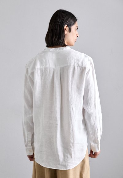 Camicia bianca in lino con maniche lunghe, cucitura a yoke sul retro e polsini con bottoni, esposta su uno sfondo neutro.