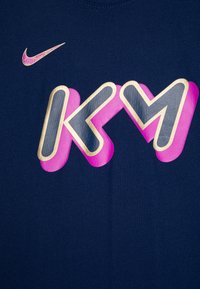 Námořní modré tričko s polstrovanou grafikou "KM" v růžové a šedé barvě. Malé zlaté logo Nike se nachází nad designem. Hladká bavlněná textura.
