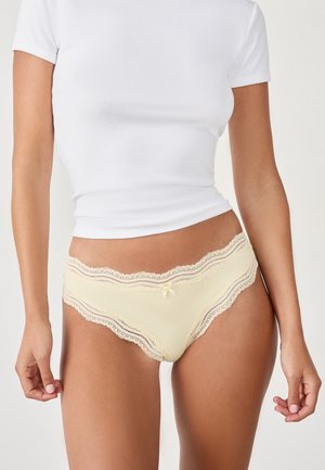 Helle gelbe Slips mit Spitzendetails und gewelltem Rand sowie einer kleinen Schleifenapplikation. Aus weichem Stoff gefertigt, mit filigranen Spitzelementen.