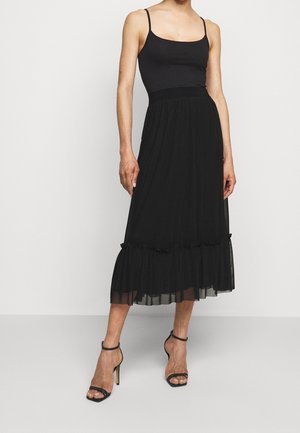 Woman wearing a black sleeveless top and a black midi skirt with ruffled hem, paired with black high-heeled sandals.