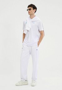 Biały strój sportowy składający się z koszulki polo i spodni dresowych, oba z niebieskimi akcentami. Model nosi białe sneakersy i okulary przeciwsłoneczne, a na ramionach ma zarzucony sweter.