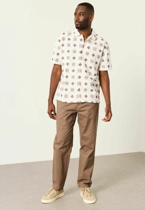 Mann trägt beige gemustertes Kurzarmhemd, braune Hose und beige Sneakers, steht und blickt nach links vor einem schlichten Hintergrund.