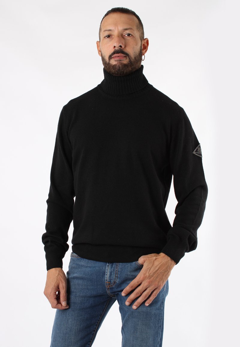Maglione a collo alto nero realizzato in morbido tessuto, con colletto e polsini a coste, e un logo applicato sul maniche sinistro. Indossato con jeans blu.