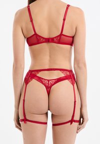 Vrouw in een rode kanten lingerieset met beha, haltergordel, string en bovenbeenbanden, van achteren zichtbaar op een witte achtergrond.